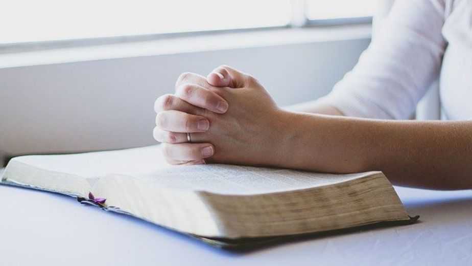 a woman praying with hands folded on a desk with open bible