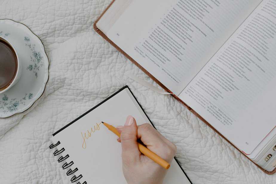 On a desk there is an open bible, coffee, and an open notebook with a woman's hand writing "Jesus" on it.