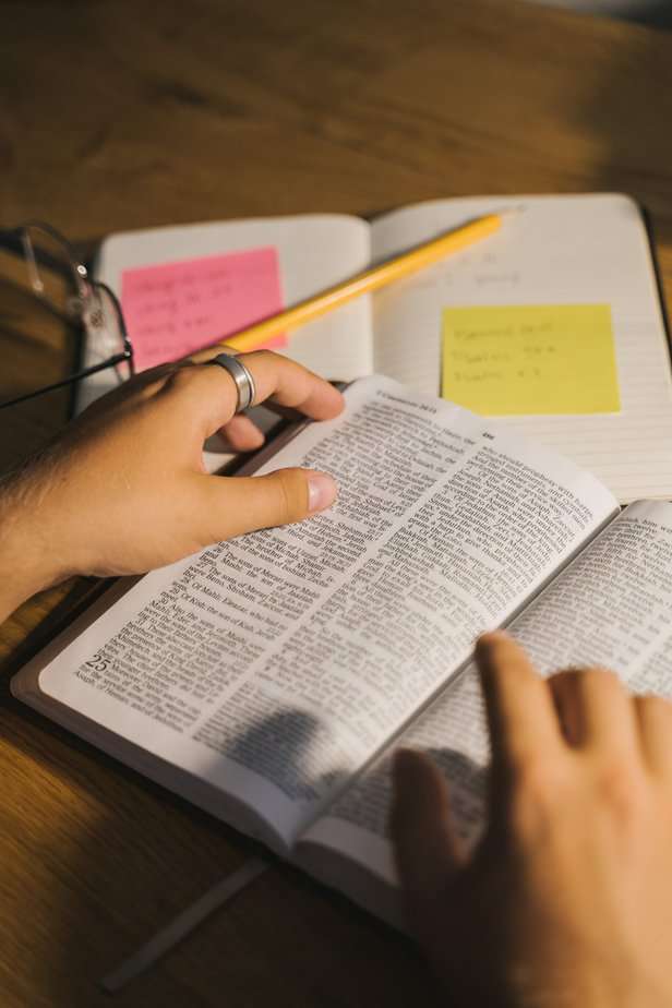 Open bible on the desk with hands flicking through the pages and open notebook with postit notes and pencil for note making on the side with glasses to read