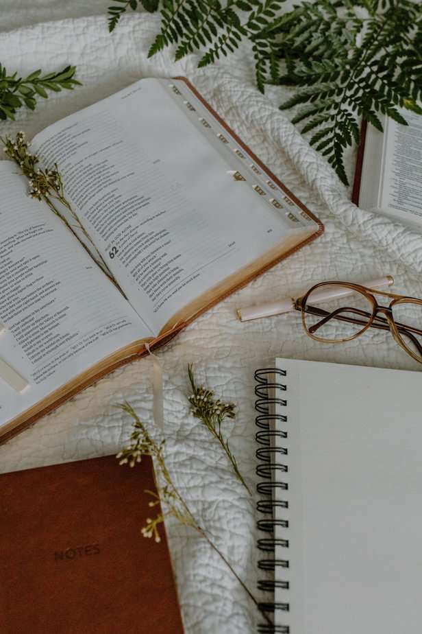 Open Bible on the desk with glasses, notebook, and small plant leaves scattered on the table