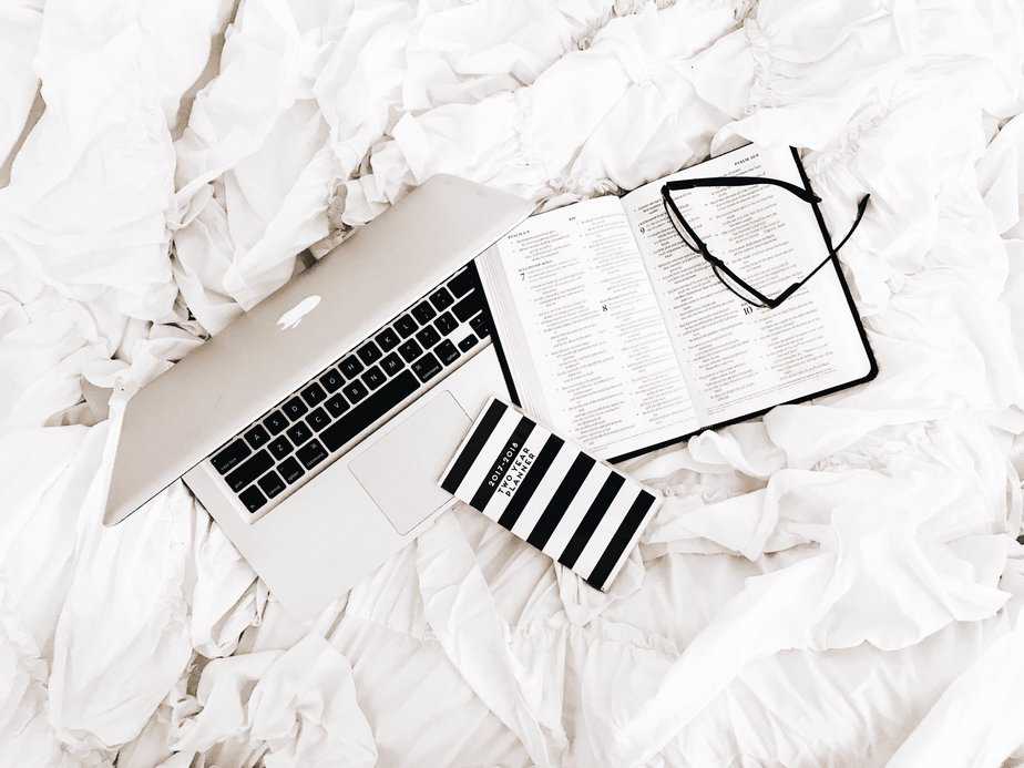 Open Bible, laptop , and glasses on a white crumbled bedsheet.